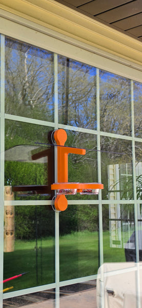 An orange recycled plastic window mounted bird feeder with a clear acrylic roof, holds two clear dishes with grape jelly.