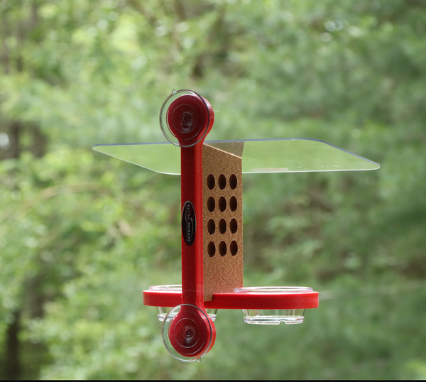 Recycled plastic window mount feeder with 2 suction cups and red mounting bracket. Has 12 holes on both sides of brown body, a clear acrylic roof, and red base holds 2 clear cups. 