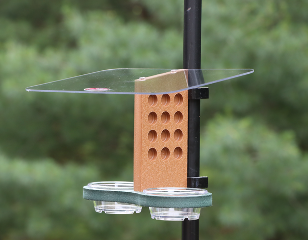 A recycled plastic bird feeder clips to a black metal pole outdoors, featuring a clear acrylic roof, a brown vertical piece with circular holes, and a green tray with two small clear bowls.