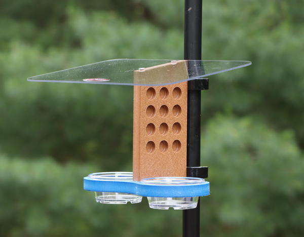 A recycled plastic bird feeder clips to a black metal pole outdoors, featuring a clear acrylic roof, a brown vertical piece with circular holes, and a blue tray with two small clear bowls.