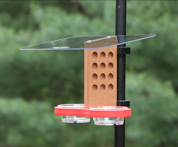 A recycled plastic bird feeder clips to a black metal pole outdoors, featuring a clear acrylic roof, a brown vertical piece with circular holes, and a red tray with two small clear bowls.