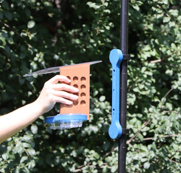 A hand holds a brown and blue bird feeder, with two clear cups and a clear sloped roof, a few inches away from a blue mounting bracket attached to a black pole with two clips. 