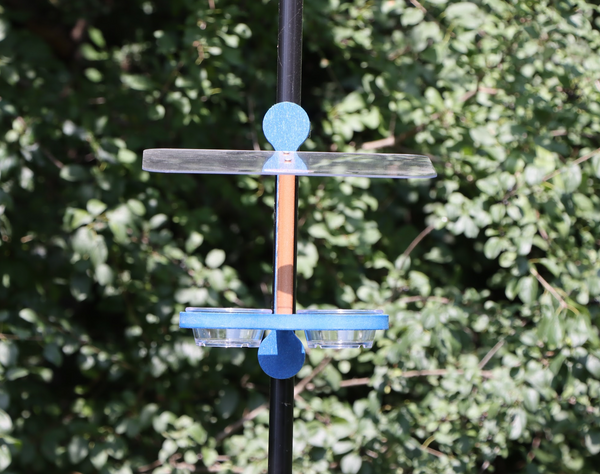 Straight on view of a bird feeder clipped to a black pole featuring a brown center, a blue mounting bracket and base, black plastic clips, a clear roof, and two small clear bowls, set against a background of green foliage.