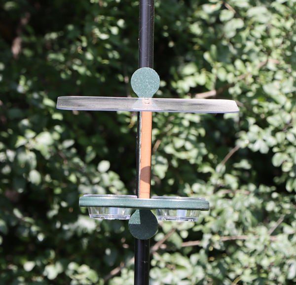 Straight on view of a bird feeder clipped to a black pole featuring a brown center, a green mounting bracket and base, black plastic clips, a clear roof, and two small clear bowls, set against a background of green foliage.