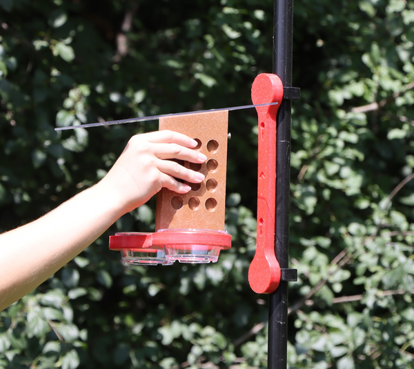A hand holds a brown and red bird feeder, with two clear cups and a clear sloped roof, a few inches away from a blue mounting bracket attached to a black pole with two clips. 