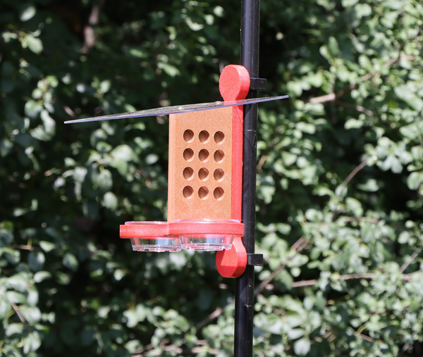 A bird feeder clipped to a black pole, featuring a brown center with circular holes, a red mounting bracket and base, black plastic clips, a clear roof and two small clear bowls, set against a background of green foliage.